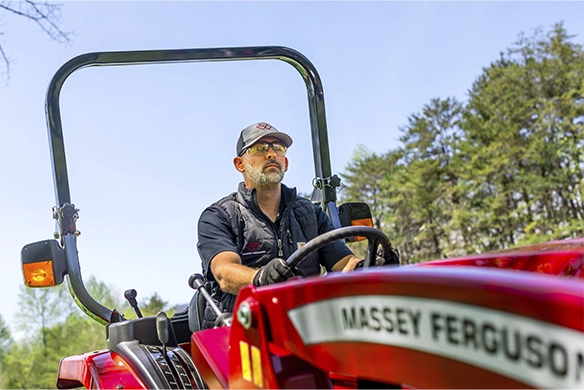 Man driving a red Massey Ferguson compact tractor through a rural area, wearing sunglasses and a cap, with trees in the background.